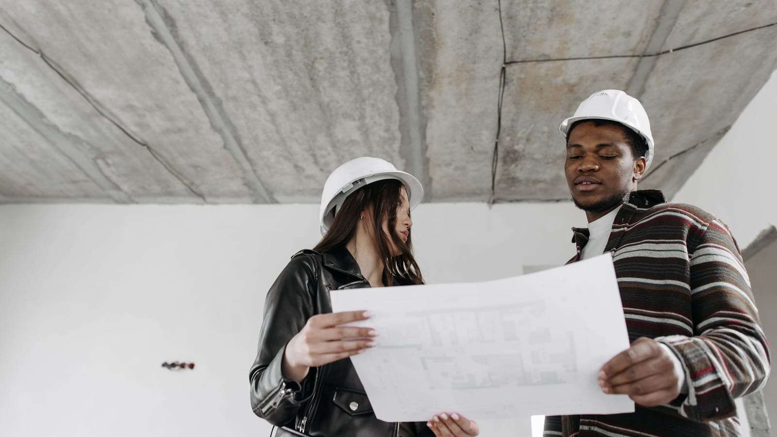 مهندسة ومهندس بخوذتين بيضاوين يراجعان مخططاً داخل مبنى قيد الإنشاء