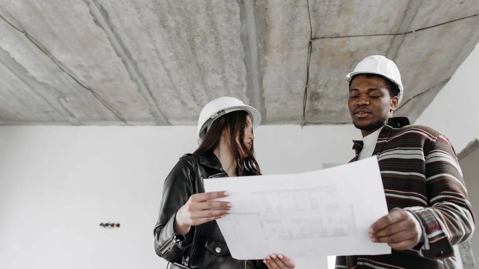 مهندسة ومهندس بخوذتين بيضاوين يراجعان مخططاً داخل مبنى قيد الإنشاء