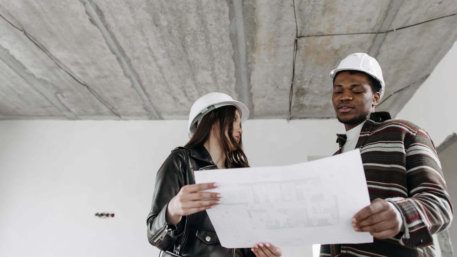 مهندسة ومهندس بخوذتين بيضاوين يراجعان مخططاً داخل مبنى قيد الإنشاء